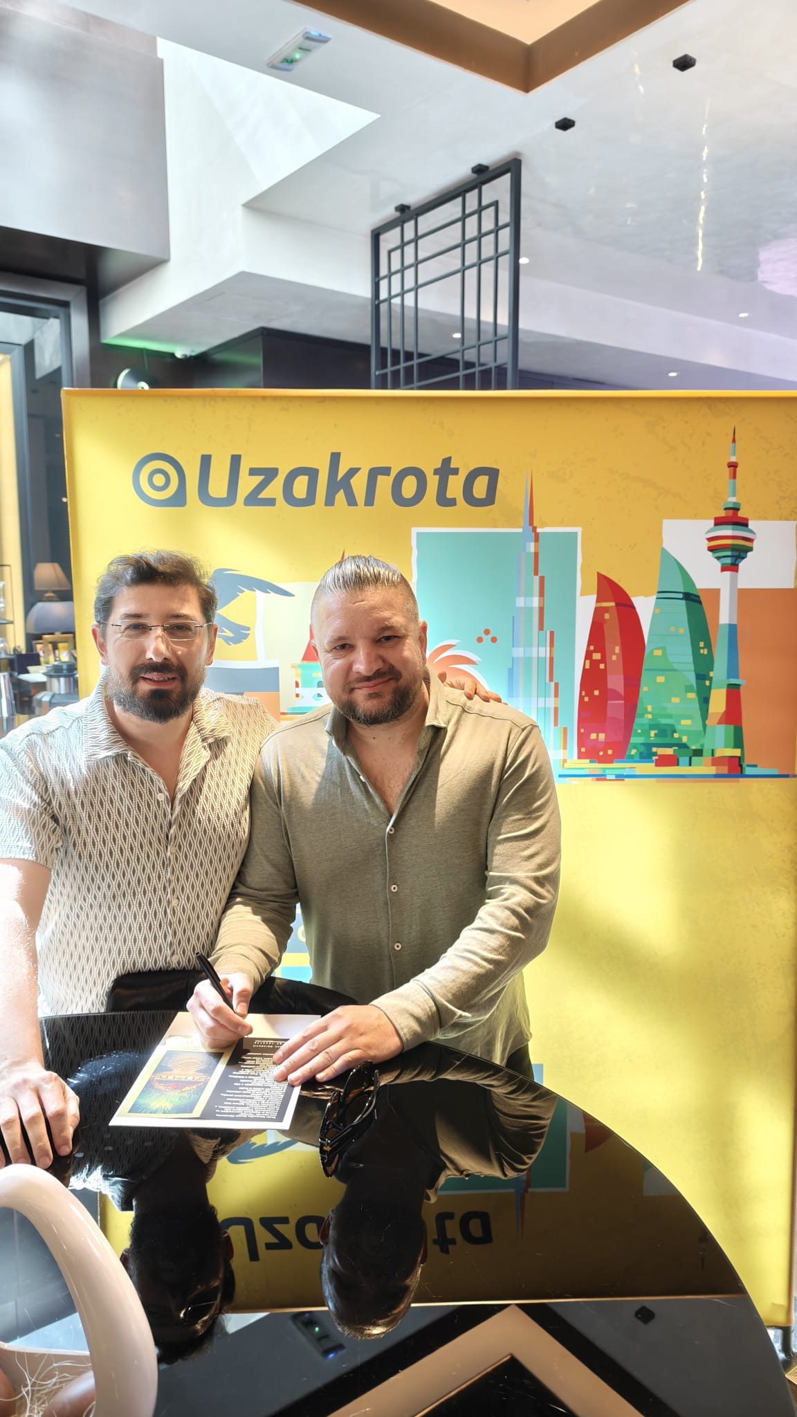 Two men pose together at a signing event in front of a bright yellow Uzakhrota banner with a colorful cityscape illustration on it.