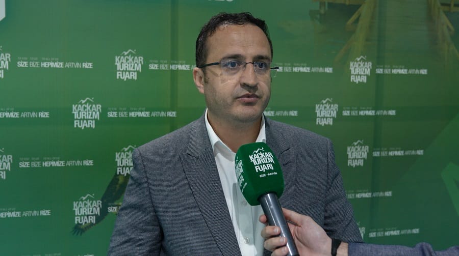 Man in a gray blazer being interviewed at a green tourism fair backdrop, holding a green microphone.