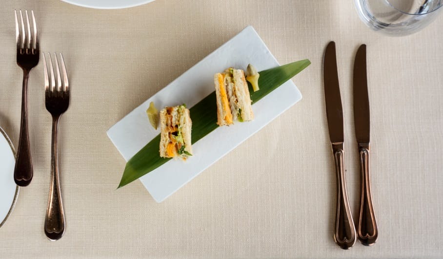 Two bite-sized sandwich canapés on a green leaf atop a white rectangular plate, with copperware on a beige table setting.