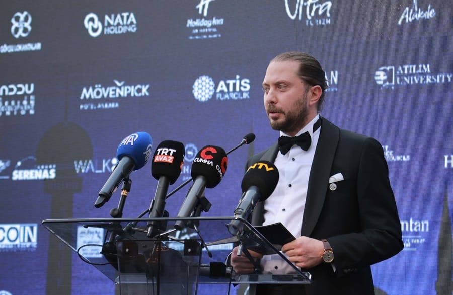 Man in a black tuxedo giving a speech at a clear podium with multiple microphones in front, backdrop covered with logos.