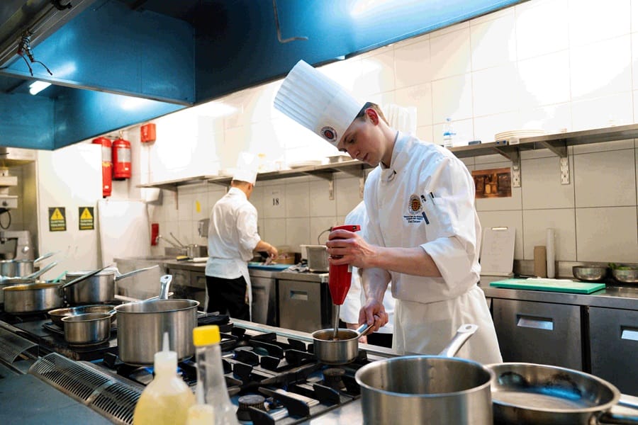 Chef in white uniform and tall hat uses a red squeeze bottle over a small pot on a busy commercial stove.