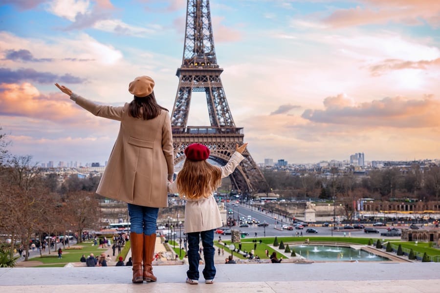 A woman and a child stand with their backs to the camera, facing the Eiffel Tower at sunset from a viewpoint.