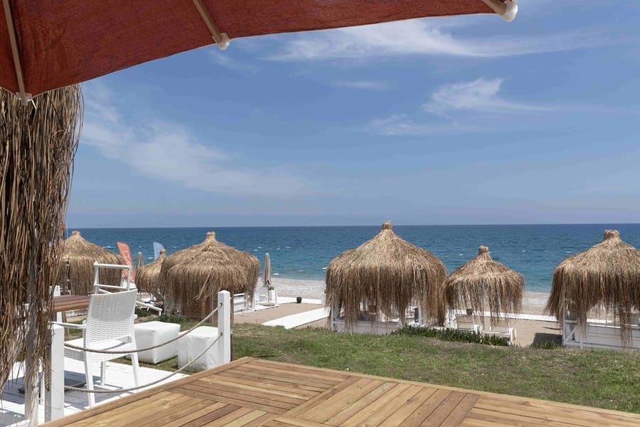 Beach scene with straw-thatched umbrellas along the shore, blue ocean, and clear sky; wooden deck furniture in foreground.