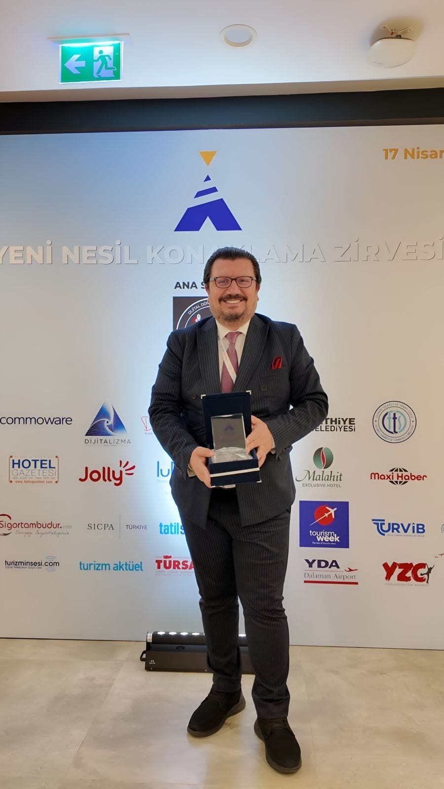 Man in a pinstripe suit holding an open award box, posing in front of a sponsor wall with logos and banners behind him.