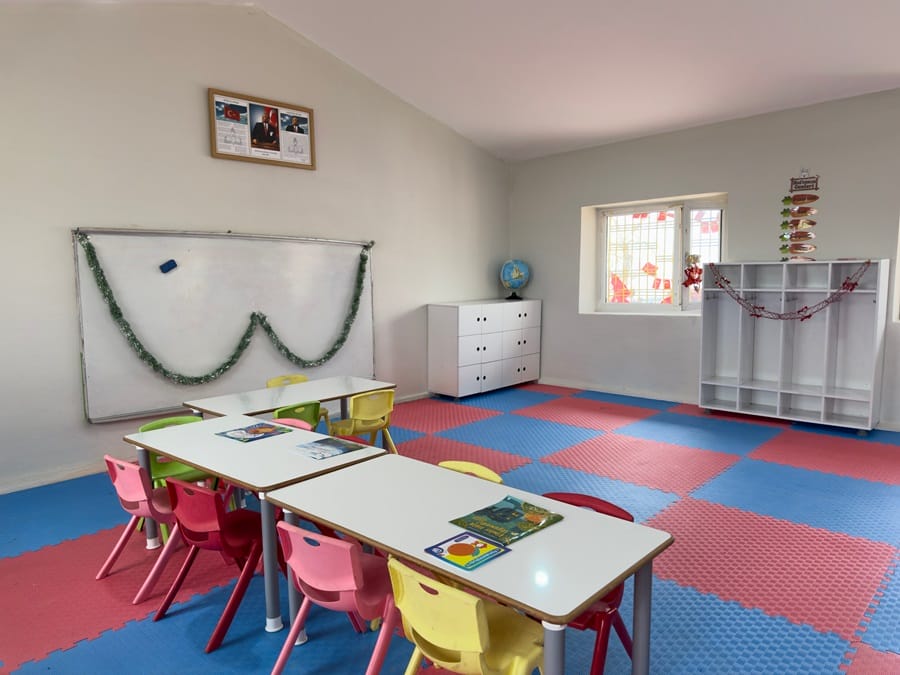 Kindergarten classroom with colorful foam mats, small tables and pink, yellow, and green chairs near a whiteboard decorated with garlands.