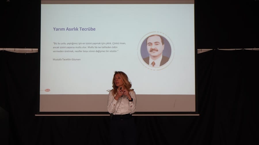 Woman on stage giving a presentation, projection slide with Turkish title 'Yarım Asırlık Tecrübe' and a portrait image on the right.